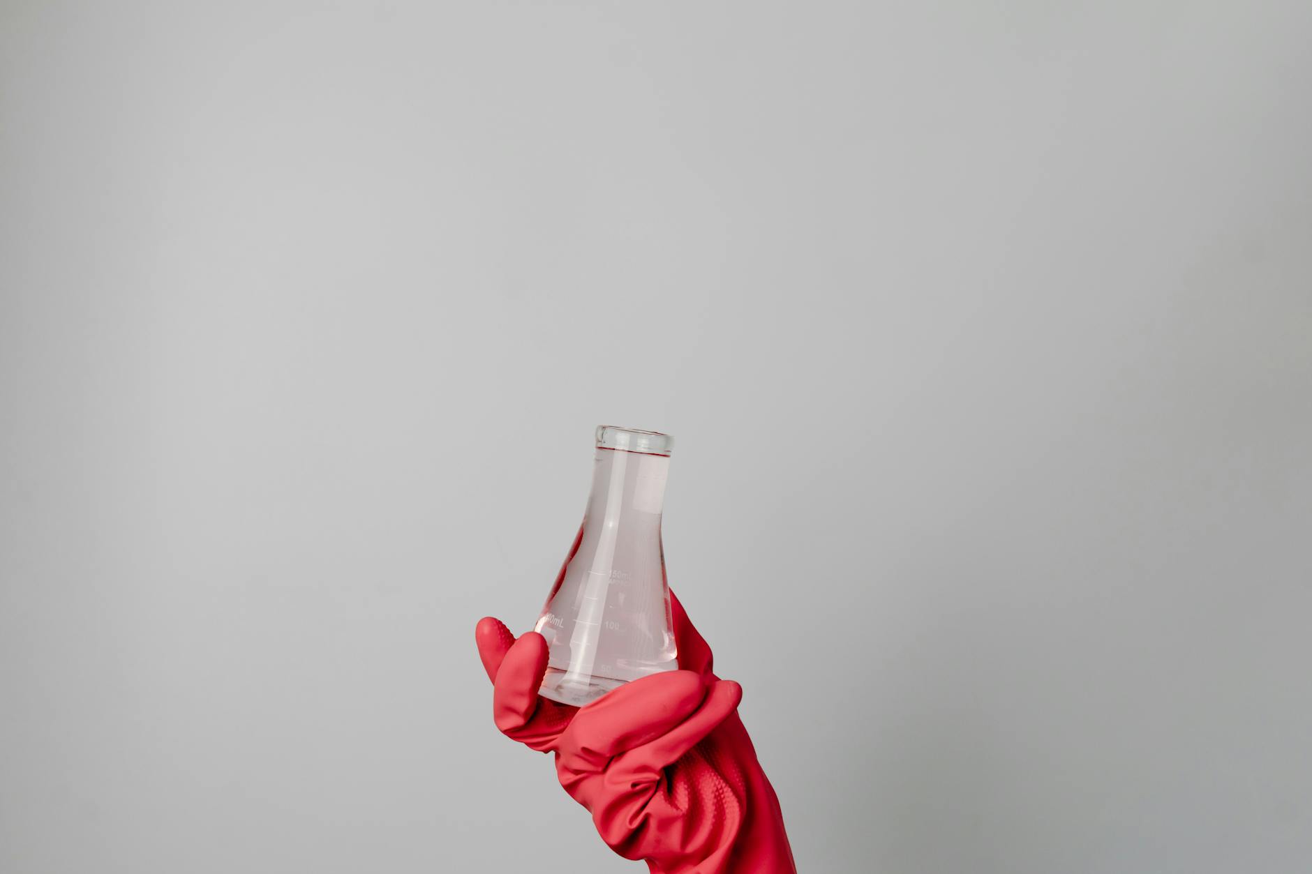A hand in a red glove holding a laboratory flask against a neutral background.