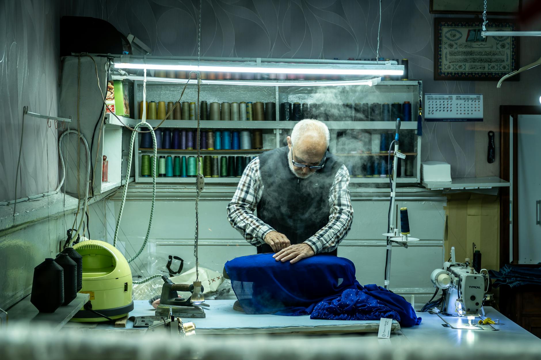An elderly tailor meticulously works with fabric in a well-equipped sewing workshop, surrounded by threads and sewing machines.