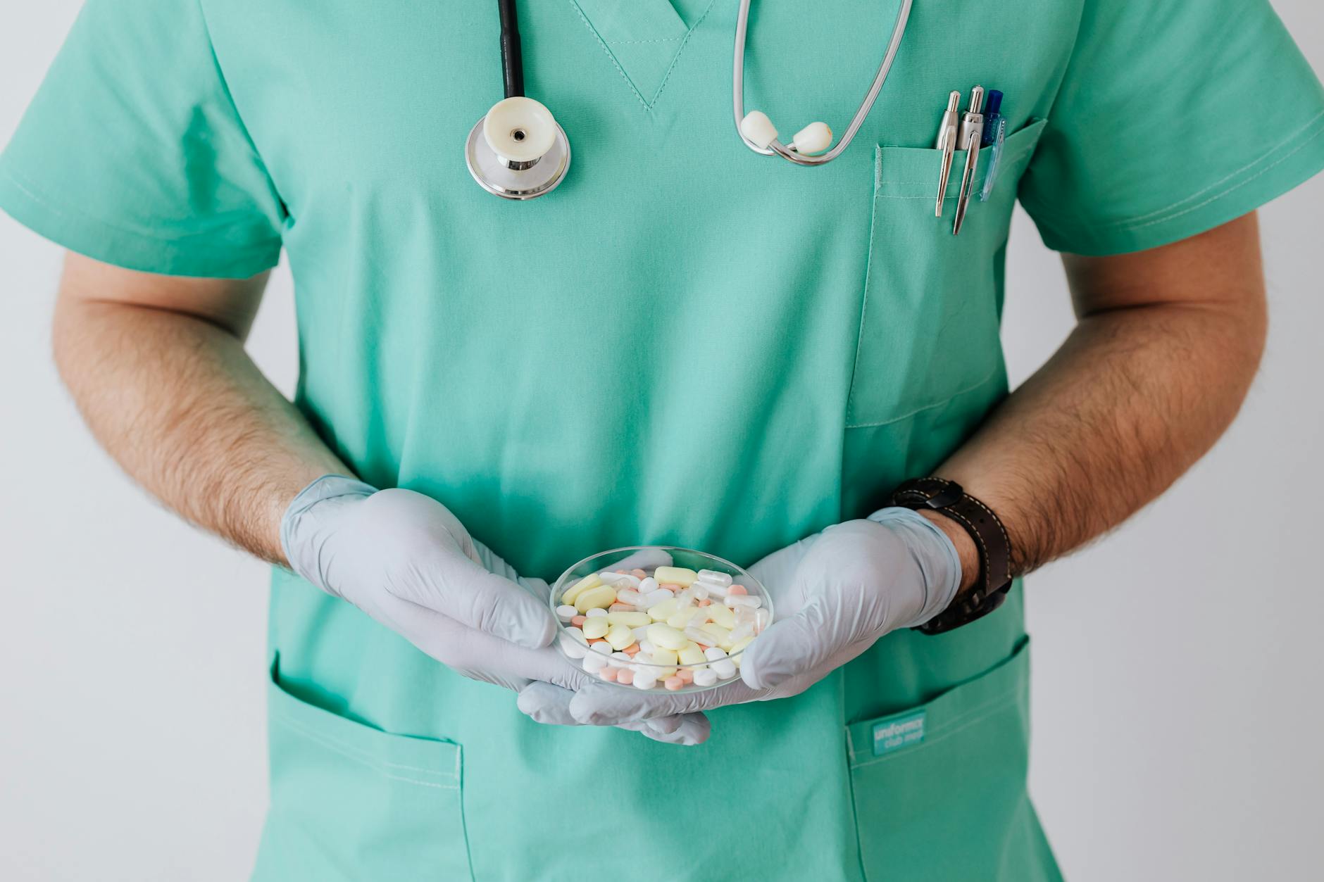 Unrecognizable male doctor wearing medical uniform and latex gloves holding Petri dish filled with assorted drugs while standing near white wall in hospital